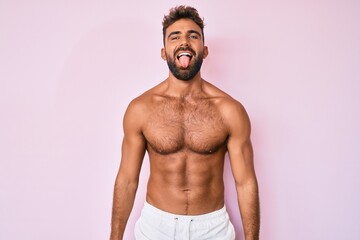 Young hispanic man standing shirtless sticking tongue out happy with funny expression. emotion...
