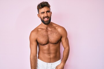 Fototapeta premium Young hispanic man standing shirtless winking looking at the camera with sexy expression, cheerful and happy face.