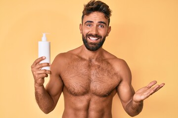 Fototapeta premium Young hispanic man standing shirtless holding sunscreen lotion celebrating achievement with happy smile and winner expression with raised hand