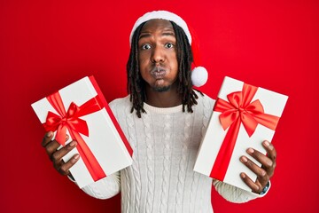 African american man with braids wearing christmas hat and holding gifts puffing cheeks with funny face. mouth inflated with air, catching air.