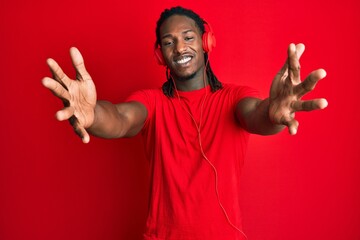 African american man with braids listening to music using headphones looking at the camera smiling with open arms for hug. cheerful expression embracing happiness.