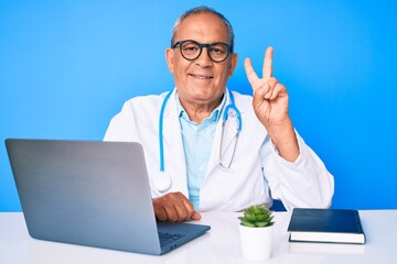 Senior handsome man with gray hair wearing doctor uniform working using computer laptop showing and pointing up with fingers number two while smiling confident and happy.