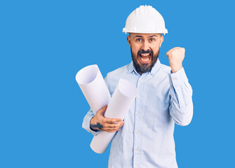 Young handsome man wearing hardhat holding paper blueprints screaming proud, celebrating victory and success very excited with raised arms