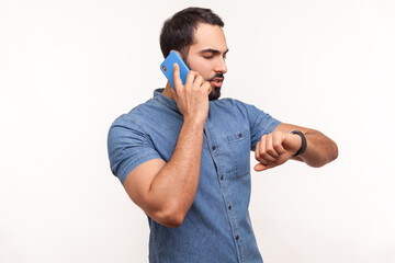 Busy bearded man in blue shirt talking phone and looking at wristwatch, making appointment, waiting...