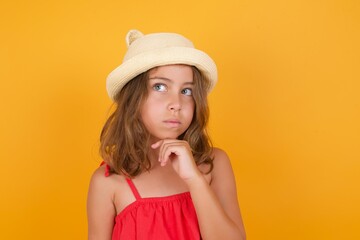 Shot of contemplative thoughtful young Caucasian girl standing against yellow background keeps hand under chin, looks thoughtfully upwards.