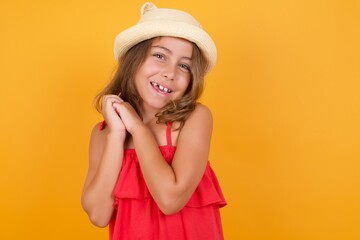 Dreamy young Caucasian girl standing against yellow background,  with pleasant expression, closes eyes, keeps hands crossed near face, thinks about something pleasant