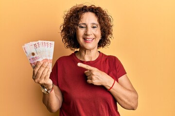 Beautiful middle age mature woman holding 10 colombian pesos banknotes smiling happy pointing with...