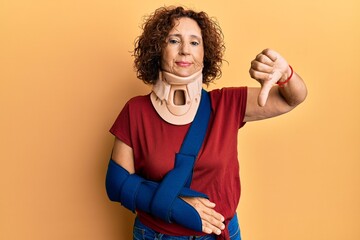 Beautiful middle age mature woman wearing cervical collar and arm on sling with angry face, negative sign showing dislike with thumbs down, rejection concept
