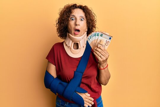 Beautiful middle age mature woman wearing cervical collar and sling holding insurance euros afraid and shocked with surprise and amazed expression, fear and excited face.
