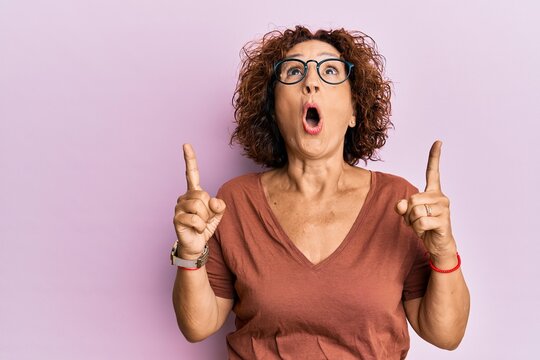 Beautiful middle age mature woman wearing casual clothes and glasses amazed and surprised looking up and pointing with fingers and raised arms.