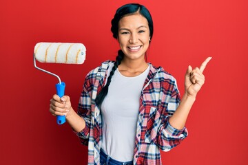 Beautiful hispanic woman holding roller painter smiling happy pointing with hand and finger to the side