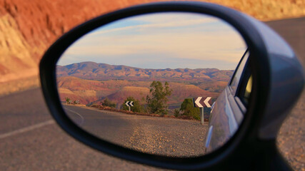 Beautifully red colored foothills of the stunning Atlas Mountains in the evening sun with paved street and road signs near Ouzoud, Morocco viewed in the side mirror of a small car. Focus on center.