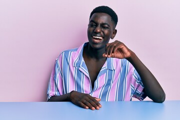 Young african american man wearing casual clothes sitting on the table winking looking at the...
