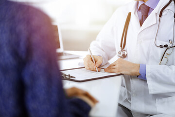 A doctor is writing a medical prescription for his patient, while sitting together at the desk in the sunny cabinet in a hospital. Physician using clipboard for filling up medication history records