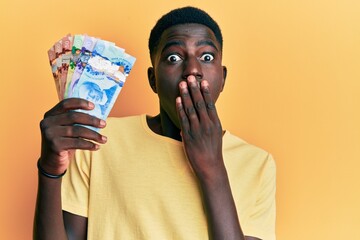 Young african american man holding canadian dollars covering mouth with hand, shocked and afraid...