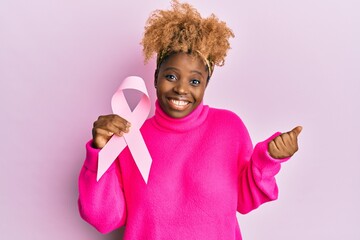 Young african woman with afro hair holding pink cancer ribbon screaming proud, celebrating victory and success very excited with raised arm