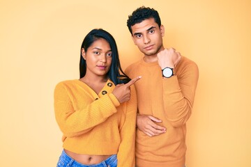Beautiful latin young couple wearing casual clothes together in hurry pointing to watch time,...