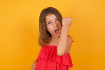 Overjoyed Caucasian young girl standing against yellow background glad to receive good news, clenching fist and making winning gesture.