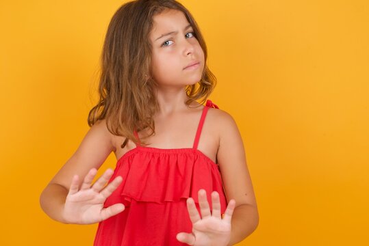 Afraid Caucasian young girl standing against yellow background, makes terrified expression and stop gesture with both hands saying: Stay there. Panic concept.