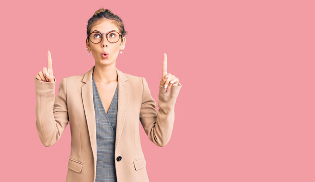 Beautiful caucasian woman with blonde hair wearing business jacket and glasses amazed and surprised looking up and pointing with fingers and raised arms.