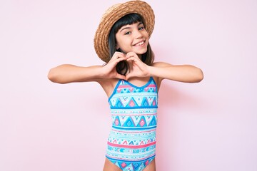 Young little girl with bang wearing swimsuit and summer hat smiling in love doing heart symbol shape with hands. romantic concept.