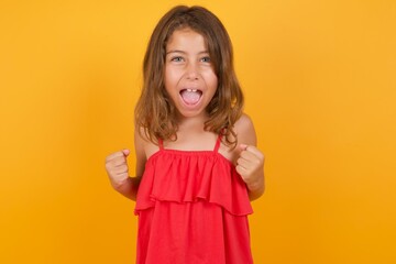 Portrait of young Caucasian girl standing against yellow background looks with excitement at camera, keeps hands raised over head, notices something unexpected reacts on sudden news.