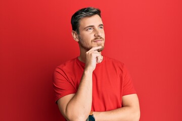 Handsome caucasian man wearing casual red tshirt with hand on chin thinking about question, pensive expression. smiling with thoughtful face. doubt concept.