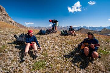 Fototapeta premium Group of trekkers resting and playing guitar in rocky pass with mountain top in high mountains. Young men and women hiking near lake Sary-Chelek Jalal Abad region, Kyrgyzstan, Trekking in Central Asia