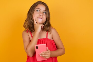 Image of a thinking dreaming Caucasian young girl standing against yellow background using mobile phone and holding hand on face. Taking decisions and social media concept.