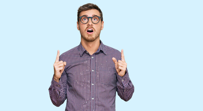 Handsome caucasian man wearing casual clothes and glasses amazed and surprised looking up and pointing with fingers and raised arms.