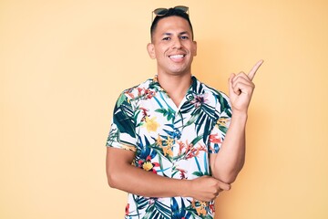 Young handsome latin man wearing summer clothes with a big smile on face, pointing with hand and finger to the side looking at the camera.