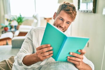 Handsome caucasian man smiling happy sitting on the sofa at home reading a book