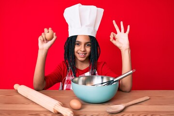 Young african american girl child with braids wearing professional cook apron holding egg doing ok...