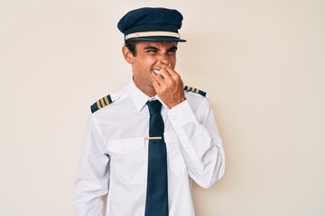 Young hispanic man wearing airplane pilot uniform smelling something stinky and disgusting, intolerable smell, holding breath with fingers on nose. bad smell