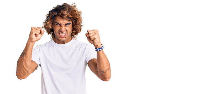 Young hispanic man wearing casual white tshirt angry and mad raising fists frustrated and furious while shouting with anger. rage and aggressive concept.