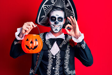 Young man wearing mexican day of the dead costume holding pumpkin doing ok sign with fingers, smiling friendly gesturing excellent symbol