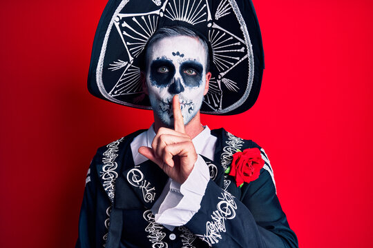Young man wearing mexican day of the dead costume over red asking to be quiet with finger on lips. silence and secret concept.