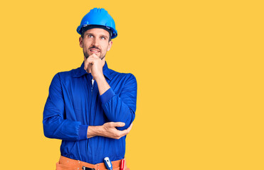 Young handsome man wearing worker uniform and hardhat with hand on chin thinking about question,...