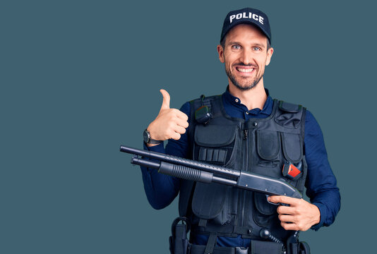 Young handsome man wearing police uniform holding shotgun smiling happy and positive, thumb up doing excellent and approval sign