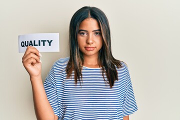 Young latin girl holding quality word on paper thinking attitude and sober expression looking self confident