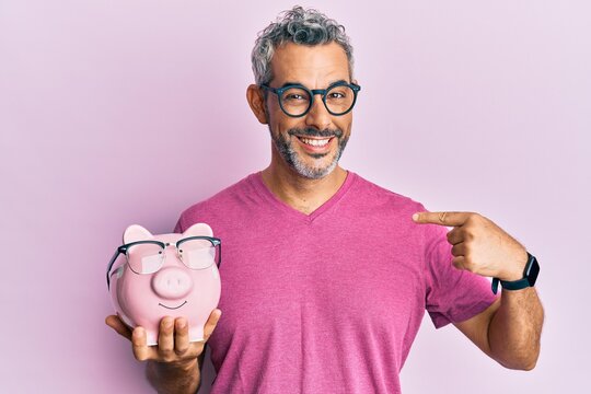 Middle age grey-haired man holding piggy bank with glasses pointing finger to one self smiling happy and proud