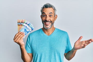 Middle age grey-haired man holding canadian dollars celebrating achievement with happy smile and...