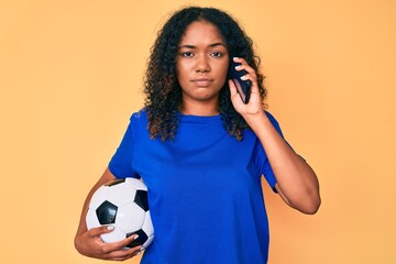 Young african american woman holding football ball talking on the smartphone relaxed with serious expression on face. simple and natural looking at the camera.