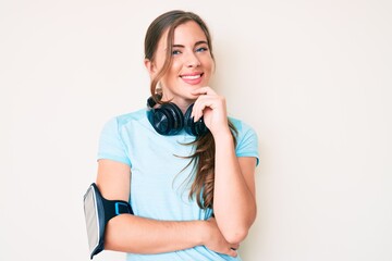 Fototapeta premium Beautiful young caucasian woman wearing gym clothes and using headphones looking confident at the camera with smile with crossed arms and hand raised on chin. thinking positive.