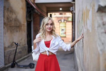 Fototapeta na wymiar Young blond woman, leaning on old building wall in city center. Three-quarter portrait of female, wearing red and white outfit, posing. Summer leisure activity. Traveler exploring the town.