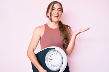 Beautiful young caucasian woman holding weight machine to balance weight loss celebrating victory...