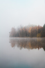 early morning view at the empty pond with the forest in the background hidden in the autumn fog and the reflection on the calm surface