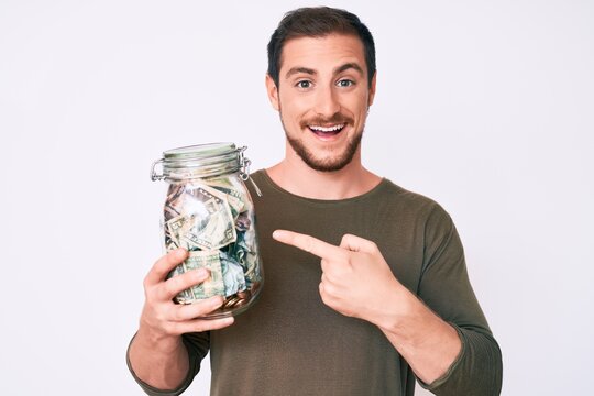 Young handsome man holding jar with savings smiling happy pointing with hand and finger
