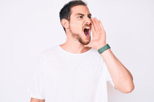 Young handsome man wearing casual white tshirt shouting and screaming loud to side with hand on mouth. communication concept.
