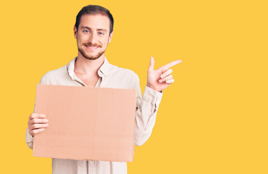 Young handsome caucasian man holding empty cardboard banner smiling happy pointing with hand and finger to the side
